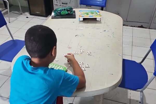 Criança de costas, sentada à mesa de uma sala de aula, monta palavras com peças de letras móveis