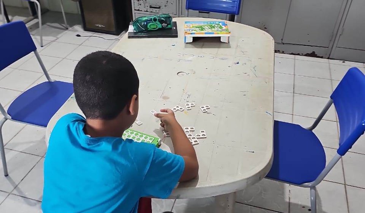 Criança de costas, sentada à mesa de uma sala de aula, monta palavras com peças de letras móveis